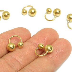 Könnte beinhalten: Goldfarbene hufeisenförmige Ohrringe mit zwei kugelförmigen Akzenten. Die Ohrringe werden auf einer Hand vor weißem Hintergrund präsentiert. Mehrere Ohrringpaare sind sichtbar.