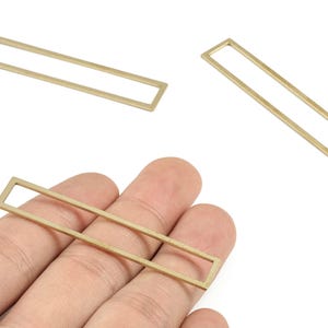 Könnte beinhalten: Drei goldfarbene, rechteckige Metall-Schmuckkomponenten. Eines wird in einer Hand gehalten, während zwei weitere in der Nähe positioniert sind. Die Komponenten haben ein einfaches, offenes Rahmendesign, das sich für die Schmuckherstellung oder das Basteln eignet.