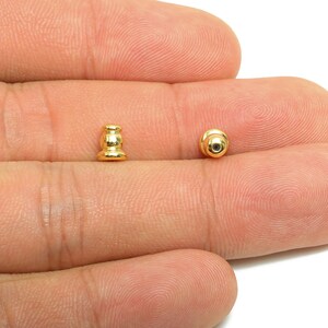 May include: Two small, gold-colored jewelry findings are displayed. One is a post with a decorative shape, and the other is a round, ball-shaped finding. The findings are shown against a skin-toned background.