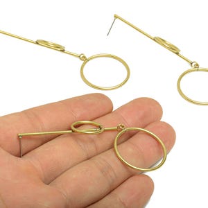 Pode incluir: Brincos geométricos dourados com um design linear. Cada brinco apresenta uma barra reta, um pequeno detalhe circular e um círculo aberto maior. Apresentados em fundo branco.