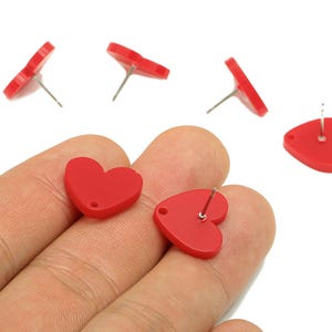 May include: Red heart-shaped stud earrings. The earrings are made of a solid red material and have a post for insertion. Several earrings are scattered on a white background, with two held in a hand.