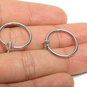 May include: Two silver hoop earring findings, each with a small, teardrop-shaped setting. The findings are held in a person's hand, showcasing the jewelry's design and potential use in crafting earrings.