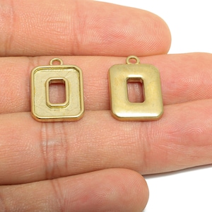 May include: Two gold-colored rectangular charms with a hollow, rectangular center. Each charm has a small loop at the top for attaching to a chain or other jewelry component. The charms are held in a hand.