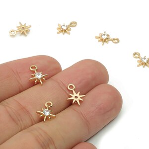 May include: Gold-colored starburst charms with a small loop for jewelry making. Some charms have a clear, round gemstone in the center. The charms are small, with a delicate design, and are shown against a white background.