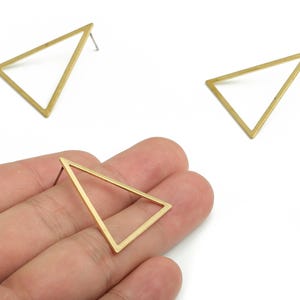 May include: Gold-colored triangle stud earrings. The earrings have a minimalist design, with a hollow triangle shape. One earring is held in a hand, while two others are on a white surface. The earrings are a geometric jewelry item.