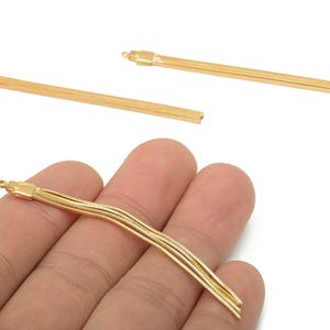 Op de afbeelding: Goudkleurige sieradenonderdelen, waaronder drie lange, dunne, rechthoekige staven en een cluster van drie kettingkwasten. De kwasten worden in een hand tegen een witte achtergrond gehouden. De staven hebben een kleine lus aan het uiteinde.