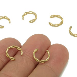 May include: A collection of gold-colored ear cuffs. The ear cuffs have a woven design. The image shows several ear cuffs scattered on a white background, with two held by a hand.