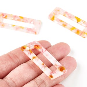 Può includere: Tre perline rettangolari in resina con un motivo marmorizzato nei toni del rosa, bianco e giallo. Ogni perlina ha un centro rettangolare cavo. Una perlina è tenuta in mano. Misure in centimetri.