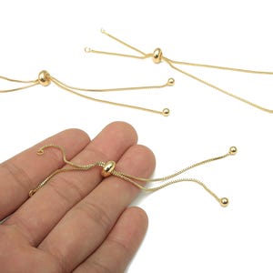 Könnte beinhalten: Goldfarbene verstellbare Armbandverschlüsse mit einer verschiebbaren Perle und zarter Kette. Jeder Verschluss hat eine kleine Kugel am Ende der Kette. Die Verschlüsse werden vor weißem Hintergrund gezeigt.