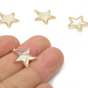 Könnte beinhalten: Goldfarbene sternförmige Anhänger mit einem perlmuttartigen Zentrum. Die Anhänger haben oben eine kleine Schlaufe zum Befestigen an Schmuck. Die Anhänger werden vor einem weißen Hintergrund gezeigt.