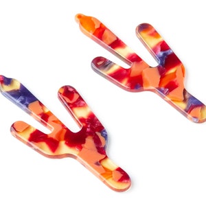 May include: A pair of colorful, abstract cactus-shaped hair clips. The clips are made of a translucent material with a swirling pattern of red, orange, yellow, and purple.