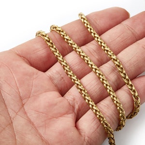 May include: Three strands of gold chain, woven in a braided pattern, are displayed on a hand.