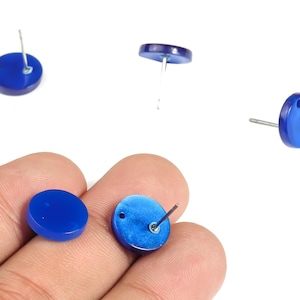 May include: A collection of cobalt blue stud earrings. The earrings are round with a post backing. One pair is held in a hand. The earrings are made of a glossy material and are set against a white background.