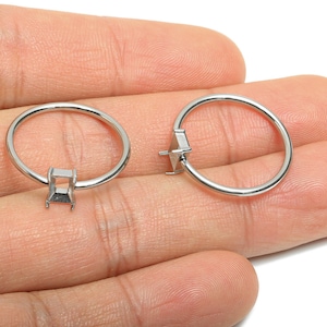 Puede incluir: Dos engastes de anillos plateados con garras cuadradas y en forma de diamante. Los anillos se sostienen en una mano, mostrando su diseño y potencial para la fabricación de joyas. Los anillos son sencillos y elegantes.