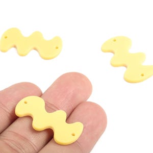 Könnte beinhalten: Drei hellgelbe, wellenförmige Acrylperlen mit jeweils zwei Löchern. Eine Perle wird in einer Hand gehalten, während die anderen beiden auf weißem Hintergrund verstreut sind. Diese Perlen eignen sich für die Schmuckherstellung und das Basteln.