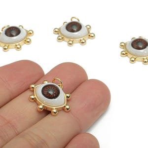 May include: Gold-colored eye-shaped pendants with a brown iris and white sclera. The pendants have a gold border with small, round accents. One pendant is held in a hand, with three others scattered nearby.