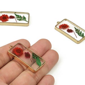 May include: Rectangular gold-toned resin pendants with dried red flowers and green leaves encased within. The pendants have a clear resin background and a gold-colored metal frame. The pendants are shown on a white background.