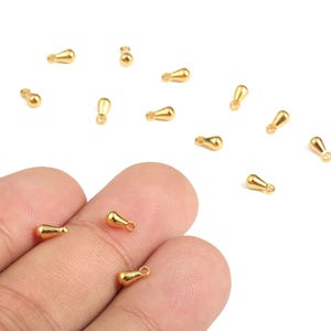 May include: Small, gold-colored teardrop-shaped jewelry findings scattered on a white surface. These findings have a loop at the top for attaching to other jewelry components. The findings are shown in a close-up shot.