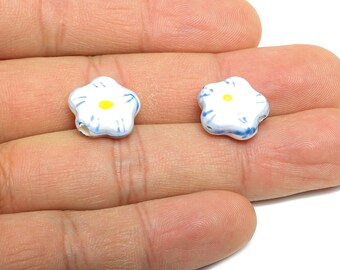Puede incluir: Dos cuentas blancas en forma de flor con centros amarillos y detalles azules. Estas cuentas de cerámica se sostienen en una mano, mostrando su delicado diseño. Ideal para la fabricación de joyas o proyectos de artesanía.