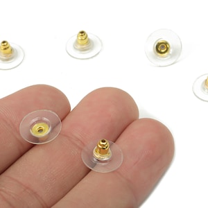 May include: A collection of earring backs. The earring backs have a clear, disc-shaped base with a gold-colored post. Several are scattered on a white background, while a few are held in a person's hand.