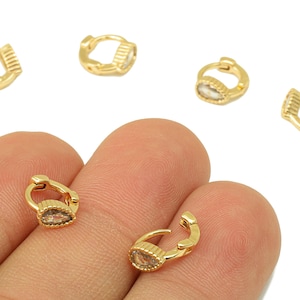 May include: Gold-colored hoop earrings with a teardrop-shaped, clear stone. The earrings are small and delicate, with a hinged closure. Several earrings are scattered on a white background, with two held by a hand.