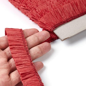 Peut inclure: Gros plan sur une frange en papier crépon rouge, dont une section est tenue dans une main. La frange est fixée à un support en carton. Le papier a un aspect texturé et froissé, et est d'une teinte rouge vif. L'image est sur fond blanc.