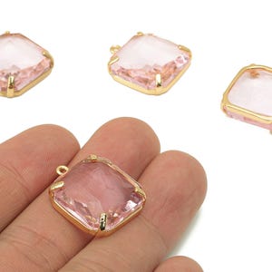May include: Four pink square-shaped crystal pendants with gold-colored metal frames. One pendant is held in a hand. The crystals have a faceted appearance, and each has a small loop for attaching to a chain or other jewelry component.