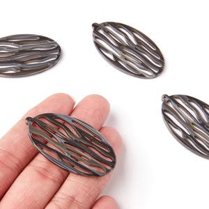 Peut inclure: Quatre pendentifs ovales en métal gris foncé avec un motif ajouré ondulé. Un pendentif est tenu dans une main. Les pendentifs ont une petite boucle en haut pour la fixation. Les pendentifs sont sur fond blanc.