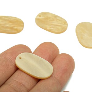 May include: Four oval-shaped, light-yellow pendants with a pearlescent finish. One pendant is held in a hand. Each pendant has a small hole at the top for stringing. These pendants are suitable for jewelry making.