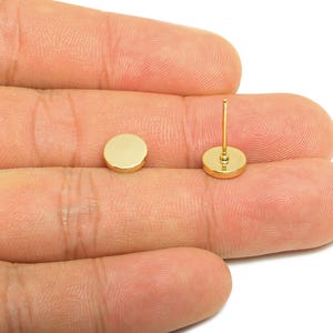 May include: Two gold-colored, round stud earrings. One earring is a flat disc, and the other has a post for insertion. The earrings are shown against a neutral background.