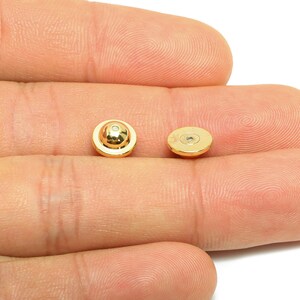 Può includere: Due piccoli elementi di gioielleria rotondi color oro. Un elemento ha un centro circolare rialzato. L'altro elemento è piatto su un lato e ha un'area incassata sull'altro lato. Gli elementi sono mostrati su uno sfondo neutro.
