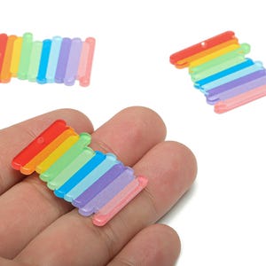 Puede incluir: Tres dijes acrílicos a rayas de arcoíris. Cada dije presenta una serie de barras paralelas en un espectro de colores, incluyendo rojo, naranja, amarillo, verde, azul, morado y rosa. Los dijes tienen un pequeño agujero para ensartar.