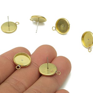 May include: Gold-colored earring posts with a flat, round base and a small loop for attaching charms. The posts have a pin for insertion into the ear. Several pieces are shown against a white background.