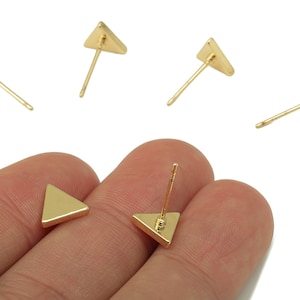 May include: Gold-colored triangle stud earrings. The earrings have a simple, geometric design with a triangular shape and a post for piercing. The earrings are shown in a close-up shot, highlighting their minimalist style and metallic finish.