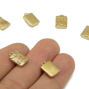 May include: Gold-colored rectangular charms with a loop at the top. Some charms have embossed floral and geometric designs. The charms are small and appear to be made of metal. The charms are shown in a hand.