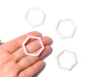 May include: Four white hexagon-shaped jewelry components with a marbled pattern. One is held in a hand. Each has a small hole for stringing. These components are suitable for crafting earrings, pendants, or other jewelry.
