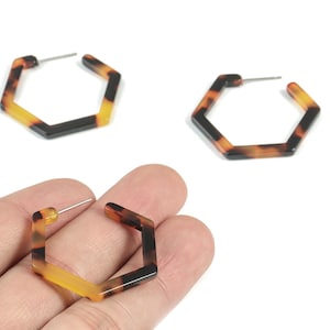 May include: Hexagon-shaped earrings in a tortoiseshell pattern. The earrings have a post for pierced ears. The color scheme includes shades of brown, black, and yellow. The earrings are shown on a white background.
