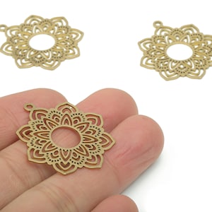 Puede incluir: Tres dijes de mandala dorados cortados con láser. Uno se sostiene en una mano, mostrando el intrincado diseño floral con un agujero central. Los otros dos están sobre un fondo blanco. Ideales para la creación de joyas.