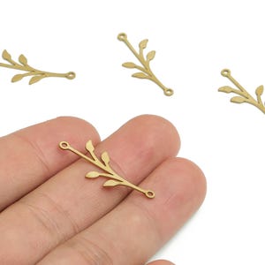 May include: Four gold-colored metal leaf branch charms. Each charm has a small loop at each end and several small leaves along the branch. One charm is held in a hand.