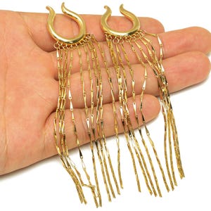 May include: A pair of gold-colored earrings with a unique design. Each earring features a curved top and multiple strands of dangling, rectangular-shaped accents. The earrings are displayed against a neutral background.