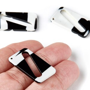 May include: Rectangular black and white resin beads with a diagonal cut-out design. The beads are shown in a close-up shot, with one held in a hand. The beads have a glossy finish and a hole for stringing.