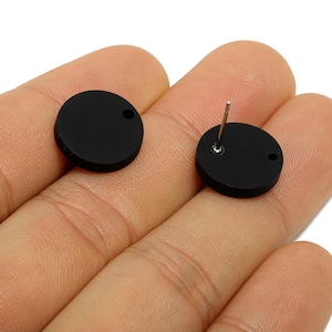 May include: Two black, circular stud earrings. One earring has a post for piercing, and the other has a small hole. The earrings are displayed on a hand.