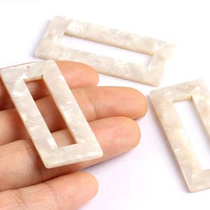 Op de afbeelding: Rechthoekige, ivoorkleurige harskralen met een open midden. De kralen hebben een gemarmerd effect en zijn ongeveer 4 cm lang en 2 cm breed. Geschikt voor het maken van sieraden en knutselprojecten.