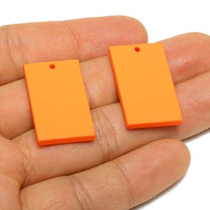 May include: Two bright orange rectangular pendants with a small hole at the top for stringing. The pendants are smooth and flat, and are held in a person's hand. The background is white.