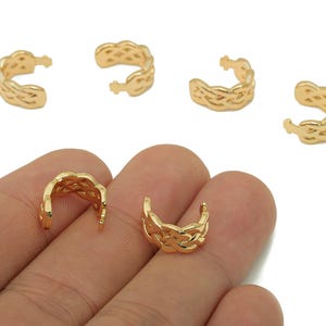 Puede incluir: Puños de oreja dorados con un diseño tejido. Los puños de oreja están en varias posiciones, algunos en una mano, otros dispersos. La joyería está hecha de un material metálico brillante, y el diseño es intrincado y detallado.