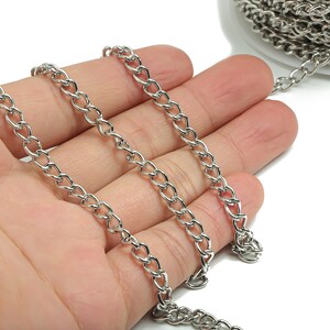 May include: A silver curb chain, a type of chain with interlocking links, is shown in a hand. The chain is laid out in a straight line, with the ends of the chain visible.