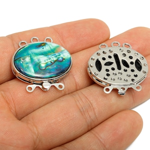 May include: Two oval-shaped jewelry clasps. One clasp features a vibrant, iridescent abalone shell inlay set in a silver-toned metal frame. The other clasp is silver-toned with a decorative openwork design. Both clasps have loops for attachment.