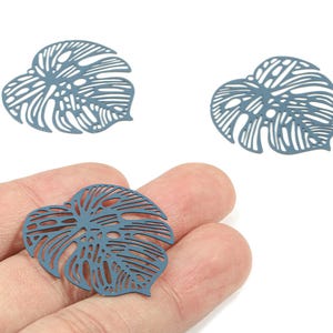 Peut inclure: Trois breloques en métal bleu-gris en forme de feuille avec des détails découpés complexes. Une breloque est tenue dans une main, tandis que deux autres sont positionnées au-dessus. Les breloques sont conçues pour ressembler à des feuilles de Monstera.