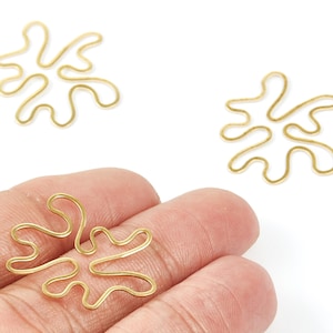 Puede incluir: Amuletos dorados con forma abstracta. Los amuletos tienen un diseño fluido y orgánico, que se asemeja a una salpicadura o ameba estilizada. Los amuletos se muestran sobre un fondo blanco, con un amuleto en una mano.