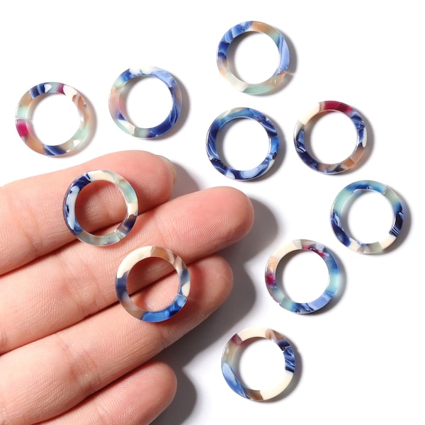 Op de afbeelding: Een verzameling kleurrijke, cirkelvormige kralen met een gemarmerd ontwerp. De kralen hebben een mix van blauwe, witte, roze en bruine tinten. Sommige kralen worden in een hand gehouden, terwijl andere op een wit oppervlak verspreid liggen. Ideaal voor het maken van sieraden.